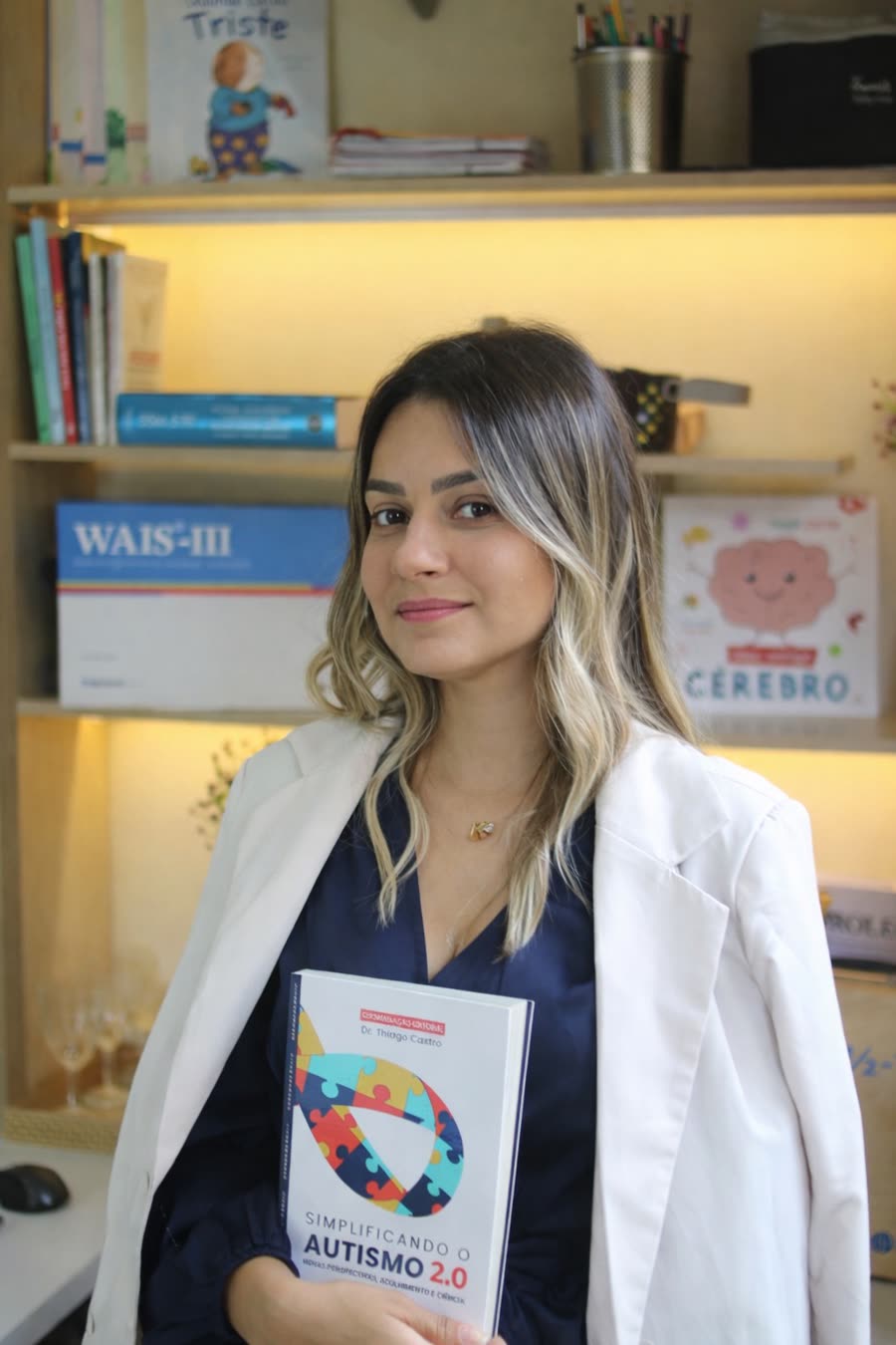 Katia Fernanda Viana de blazer branco, segurando um livro sobre autismo no consultório