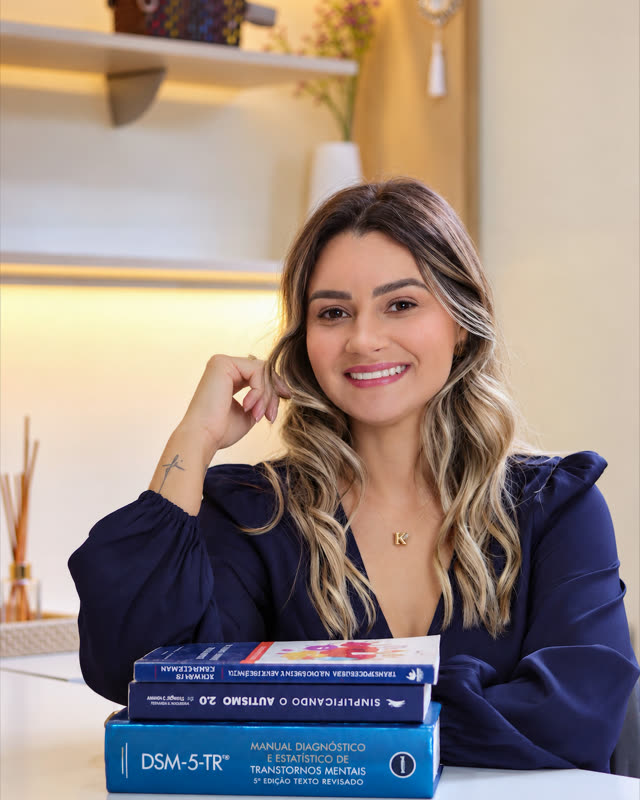 Katia Fernanda Viana sorrindo à mesa do consultório, com livros de neuropsicologia